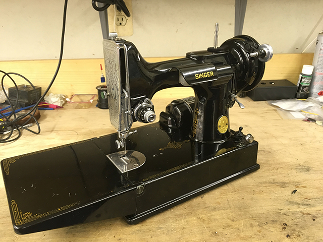 The front of a 1955 Singer Featherweight sewing machine. The machine is small, painted glossy black with decorative gold decals. 