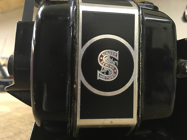 A closeup of the side of the motor showing a Singer trademark. The trademark is a red letter 'S' with the words "Singer Sewing Motors." Behind it, a woman is shown sewing at a table.